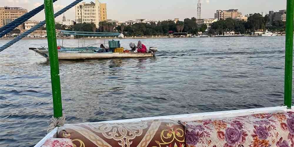 Feluca tradicional navegando pelo rio Nilo com velas brancas e paisagem natural ao fundo.