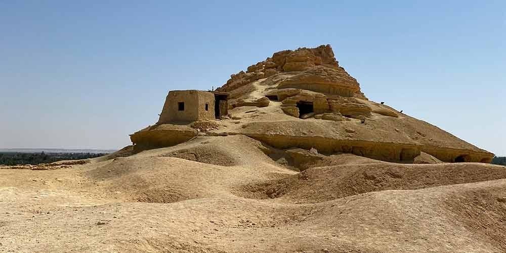 Une vue panoramique du mont des Morts (Gebel el-Mawta) à Siwa. Il s'agit d'une colline conique en pierre calcaire parsemée de nombreuses ouvertures de tombes rupestres antiques. Au sommet et sur les flancs, on voit des structures en briques de terre crue. Le sol environnant est aride et sablonneux sous un ciel bleu clair et dégagé