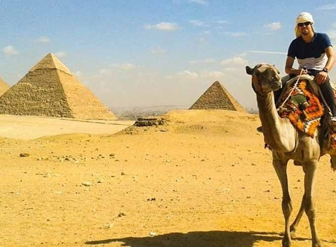A traveler riding a camel across the Giza desert with three iconic pyramids and the distant Cairo skyline visible under a bright sky. The camel wears a colorful traditional saddle, and the vast sandy landscape stretches toward the horizon during an excursion from Alexandria port