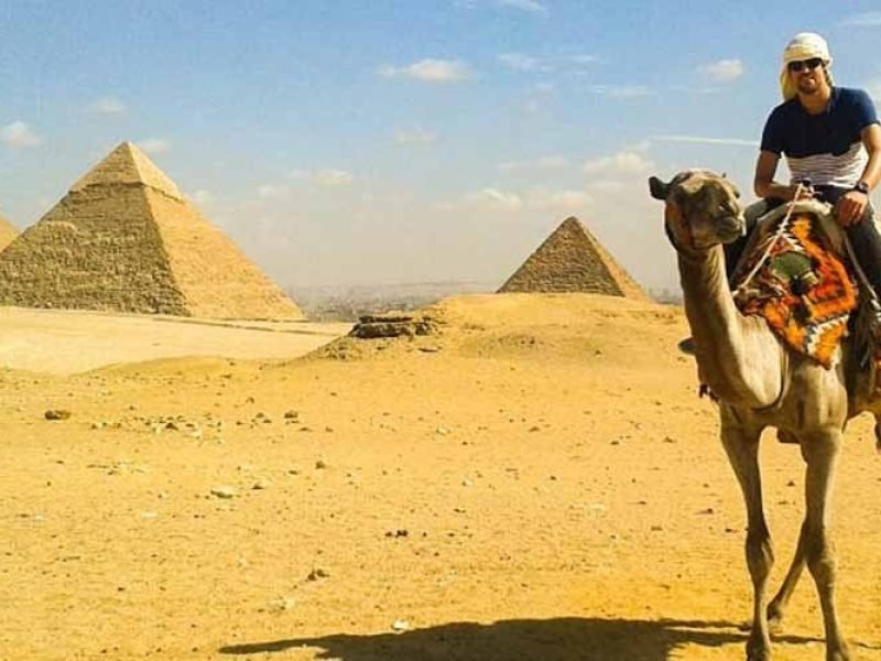 A traveler riding a camel across the Giza desert with three iconic pyramids and the distant Cairo skyline visible under a bright sky. The camel wears a colorful traditional saddle, and the vast sandy landscape stretches toward the horizon during an excursion from Alexandria port
