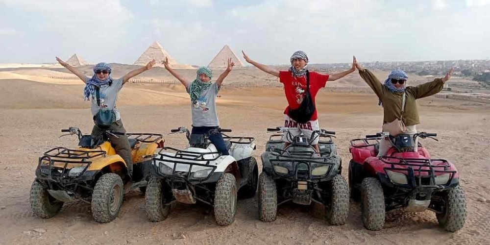 Four tourists wearing traditional headscarves pose with open arms on colorful quad bikes in the Giza desert, with the three Great Pyramids visible under a bright sky in the background