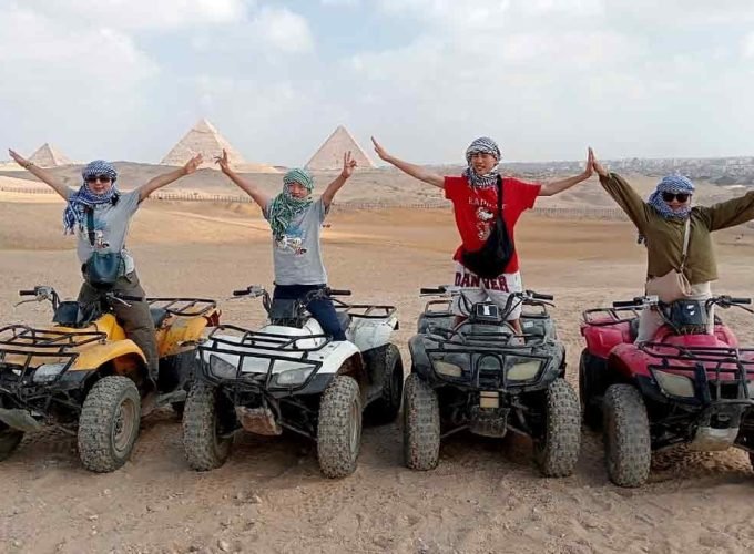 Four tourists wearing traditional headscarves pose with open arms on colorful quad bikes in the Giza desert, with the three Great Pyramids visible under a bright sky in the background