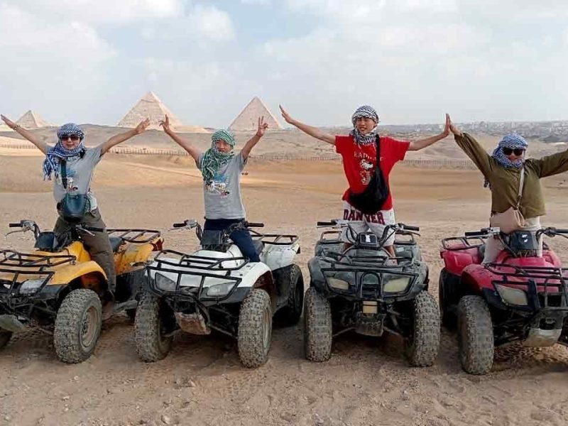Four tourists wearing traditional headscarves pose with open arms on colorful quad bikes in the Giza desert, with the three Great Pyramids visible under a bright sky in the background