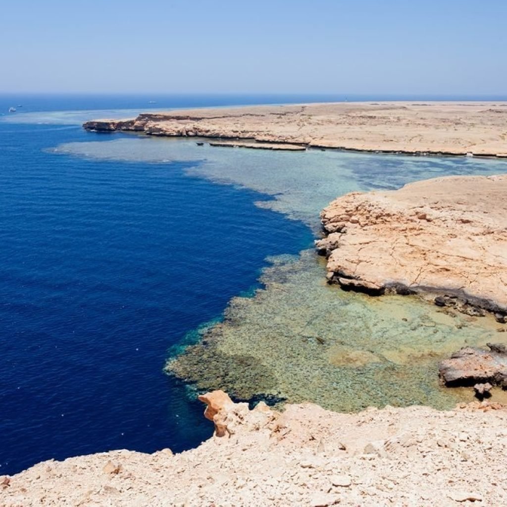 A high-angle landscape view showing the dramatic contrast between the deep blue Red Sea and the pale desert cliffs of Ras Mohammed National Park. The turquoise shallow waters reveal sprawling coral reef systems beneath the surface along the rugged, rocky coastline under a clear sky. In the far distance, small white boats are visible on the horizon of the vast, open ocean surrounding the protected Egyptian nature reserve
