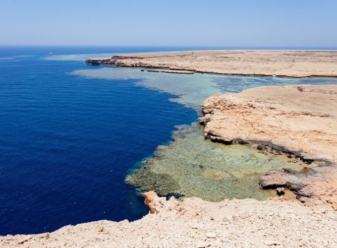 A high-angle landscape view showing the dramatic contrast between the deep blue Red Sea and the pale desert cliffs of Ras Mohammed National Park. The turquoise shallow waters reveal sprawling coral reef systems beneath the surface along the rugged, rocky coastline under a clear sky. In the far distance, small white boats are visible on the horizon of the vast, open ocean surrounding the protected Egyptian nature reserve