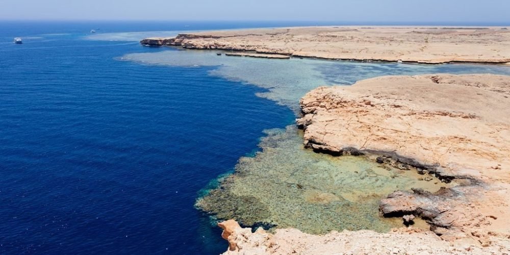 A panoramic high-angle view of the desert coastline meeting the vibrant turquoise and deep blue waters of Ras Mohammed National Park. The rugged limestone cliffs overlook a sprawling coral reef visible through the clear shallow sea under a bright, cloudless sky. Tiny white boats are seen on the distant horizon, highlighting the vastness of this protected Egyptian marine sanctuary