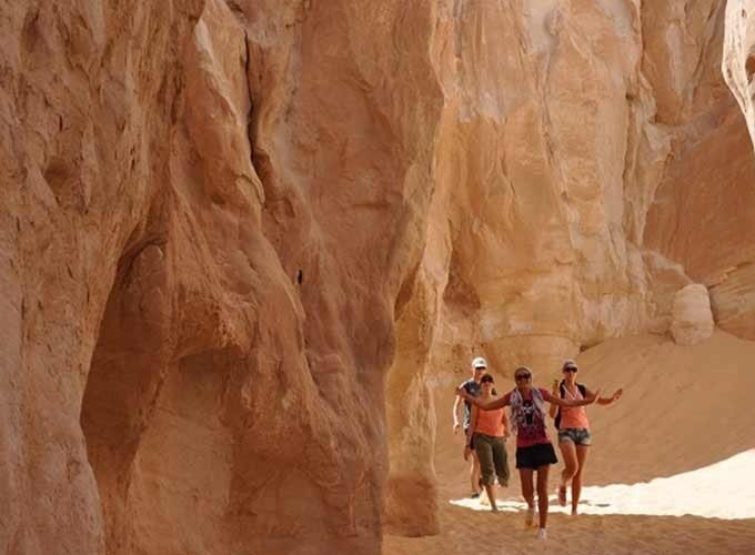 Four female tourists walking through a narrow passage between high, winding sandstone walls in a desert canyon during their safari trip. The towering rock formations feature smooth, wavy textures in shades of tan and orange, illuminated by bright natural sunlight from above. The ground is covered in soft, pale sand as the group explores the deep, natural geological corridor