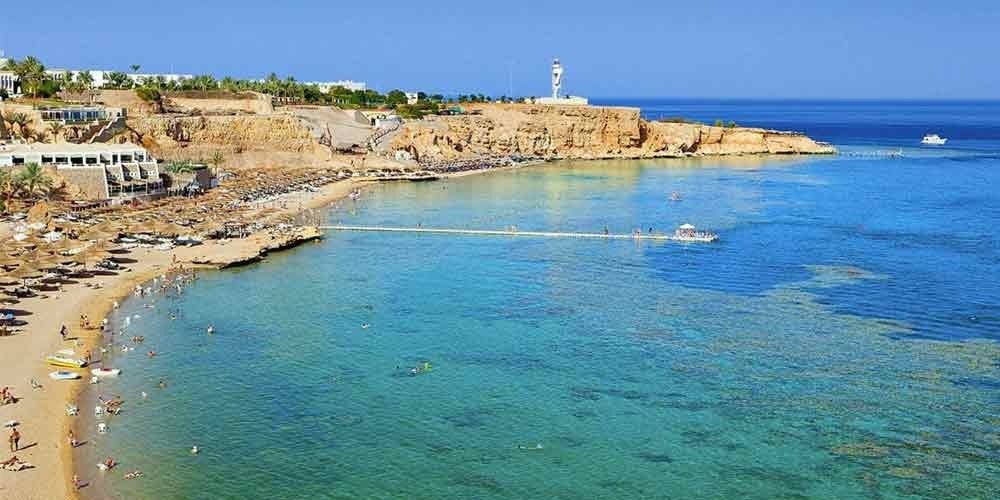 A breathtaking panoramic view of a luxury Red Sea resort featuring turquoise waters and golden sandy beaches during a Submarine trip. The coastline is dotted with sun loungers, palm trees, and a long wooden pier extending over a vibrant coral reef where people are swimming. In the distance, a white lighthouse stands atop a rugged desert cliff under a clear blue sky, overlooking a white boat sailing on the horizon