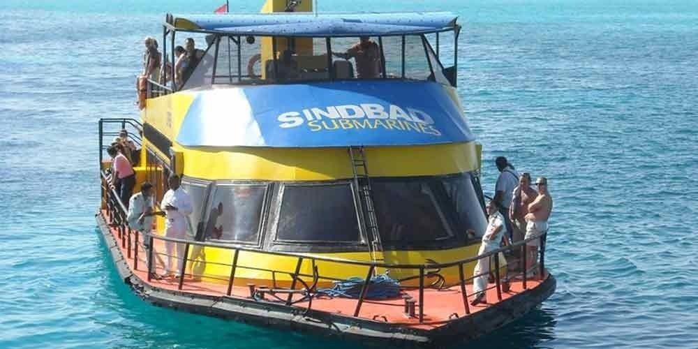 A bright yellow and blue Sindbad Submarine vessel sailing on the calm, turquoise waters of the Red Sea during a Submarine trip. The boat has a multi-level deck with tourists standing at the railing and a large panoramic glass cockpit at the front. The sun reflects off the clear blue water surrounding the vessel as it prepares for an underwater sightseeing excursion
