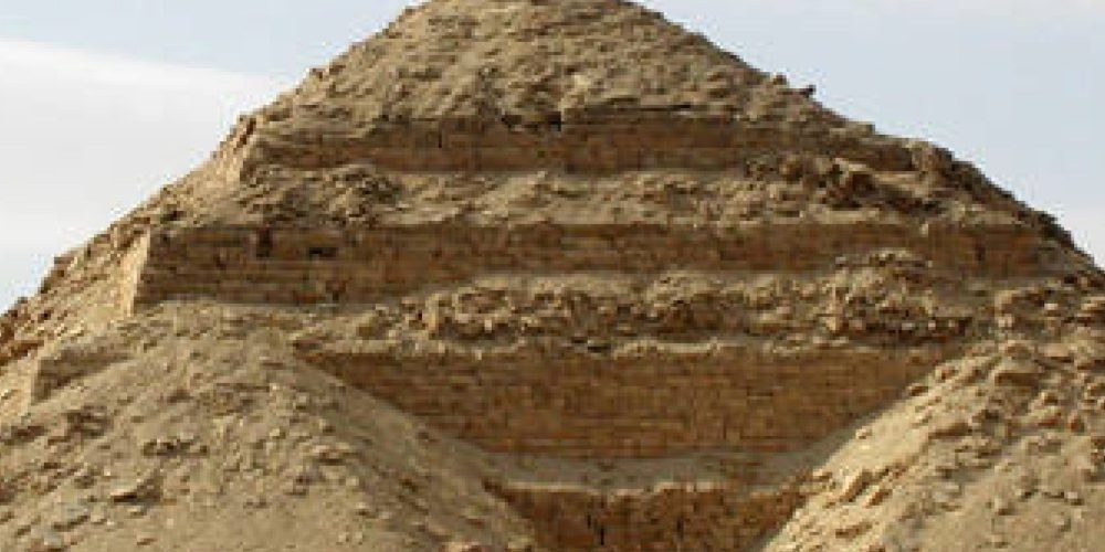 A close-up, high-angle view of a weathered ancient pyramid made of light brown stone blocks, showing a partially collapsed or unfinished top. The structure features a dark, rectangular entrance at its base, partially buried in sand. The surrounding ground is covered in fine desert sand with faint tire tracks visible on the left. The sky in the background is a pale, hazy white with light cloud cover near the Sun Temple of Niuserre