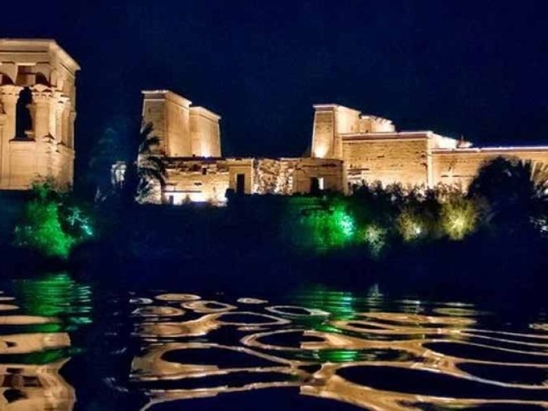 Visitantes apreciando o Templo de Philae à noite, com iluminação destacando detalhes arquitetônicos.