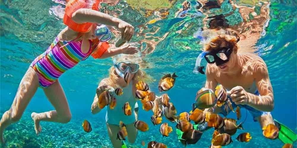 A group of three people, including two young girls in colorful swimwear, snorkeling underwater among a large school of yellow and black tropical fish. The clear turquoise water reveals a bright coral reef at the bottom during an exciting Snorkeling Trip