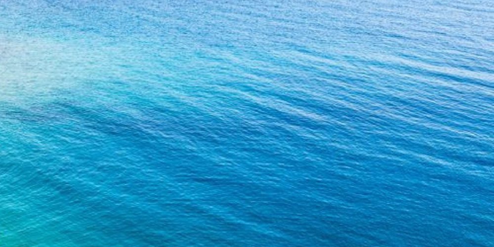 A high-angle view of a serene coastal bay with crystal-clear turquoise and deep blue water. Two boats are anchored near the shore of a desert landscape under a bright sky during a Snorkeling Trip