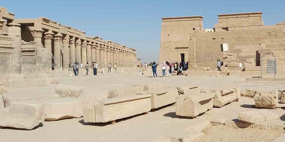 Visitors walking through an ancient Egyptian temple complex with tall stone columns and carved walls under a clear blue sky.