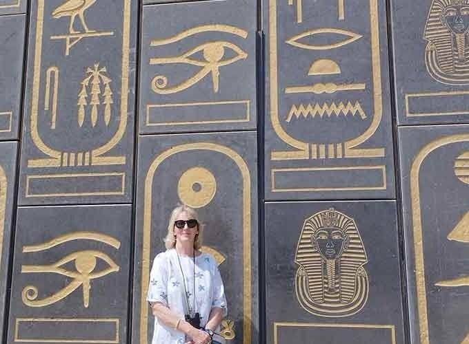 Une femme aux cheveux blonds portant des lunettes de soleil et une chemise blanche à motifs bleus pose devant un mur monumental gris foncé. Le mur est orné de grandes cartouches dorées contenant des hiéroglyphes détaillés, des représentations de pharaons et l'œil d'Horus. C'est une section impressionnante du Grand Musée égyptien