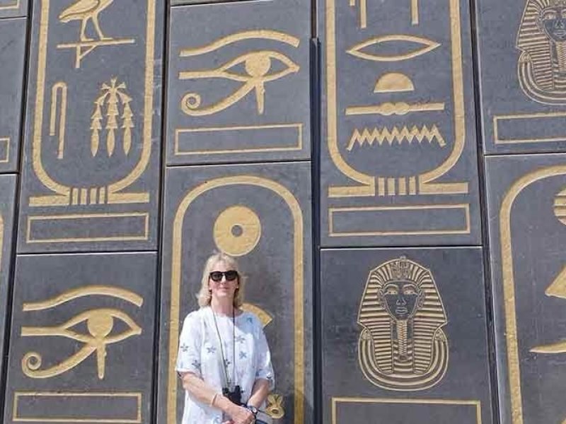 Une femme aux cheveux blonds portant des lunettes de soleil et une chemise blanche à motifs bleus pose devant un mur monumental gris foncé. Le mur est orné de grandes cartouches dorées contenant des hiéroglyphes détaillés, des représentations de pharaons et l'œil d'Horus. C'est une section impressionnante du Grand Musée égyptien