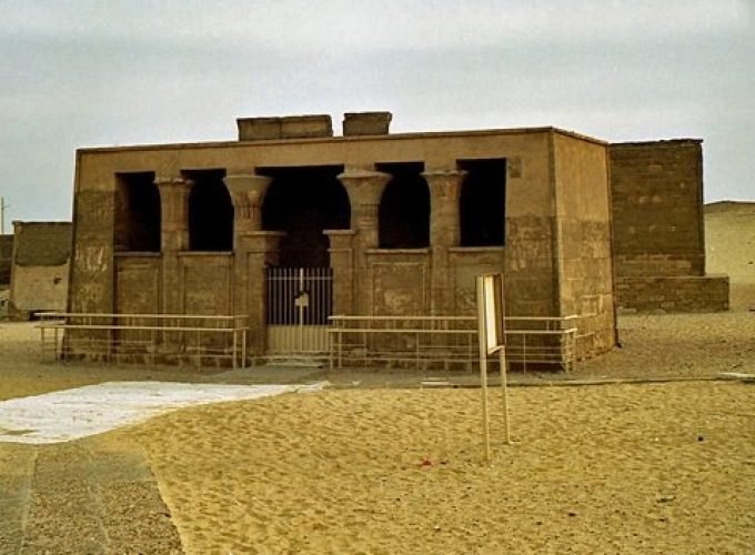 Un petit temple ou tombeau antique de forme rectangulaire en pierre calcaire beige, doté d'une entrée à colonnes sculptées derrière une grille métallique, entouré de sable fin et plat lors d'un voyage à El Minya depuis le Caire