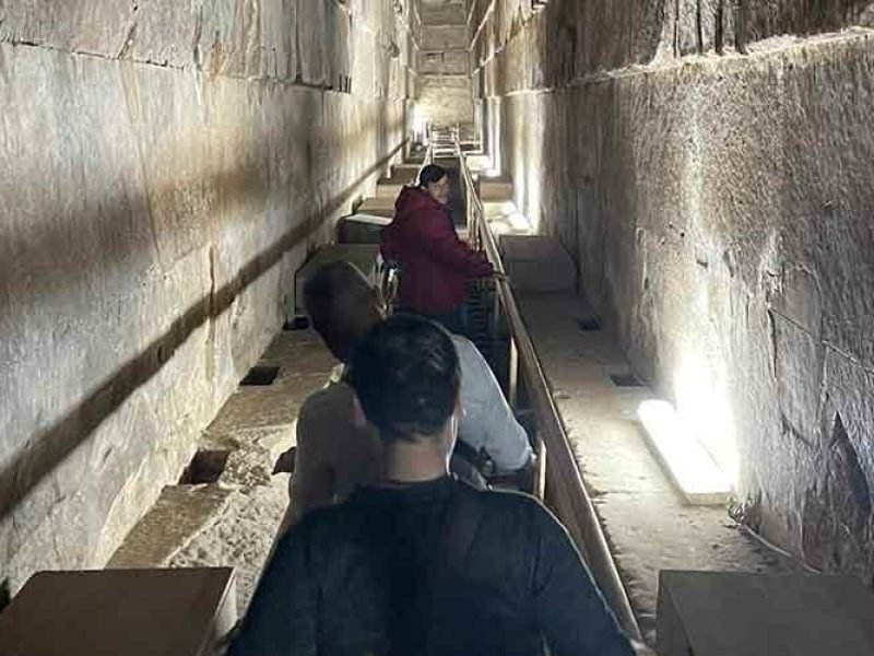 A high-angle view looking down the narrow, steep limestone corridor known as the Grand Gallery inside the Great Pyramid . Three tourists are visible: a woman in a red jacket standing further down the stairs, and two people in the foreground with their backs to the camera. The passage features tall, corbelled stone walls with a polished texture, lit by artificial floor lamps that cast long shadows. A wooden handrail runs along the side of the stepped walkway.