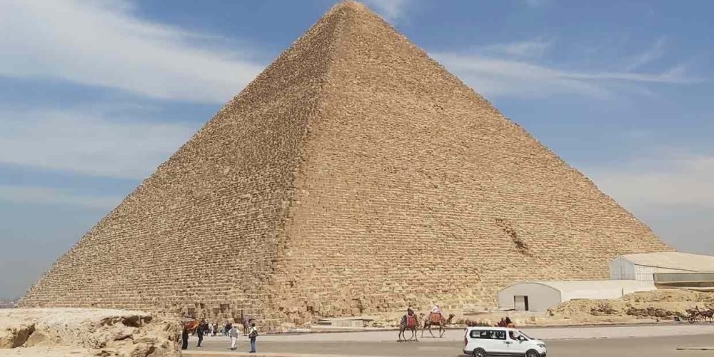 A wide exterior shot of the Great Pyramid of Giza under a clear blue sky with light wispy clouds. The massive stone structure dominates the frame. At its base, there is a paved road with a white van driving by, a few people walking, and two tourists riding camels. To the right, a long white modern building is visible. This is the starting point for a VIP tour inside the Great Pyramid
