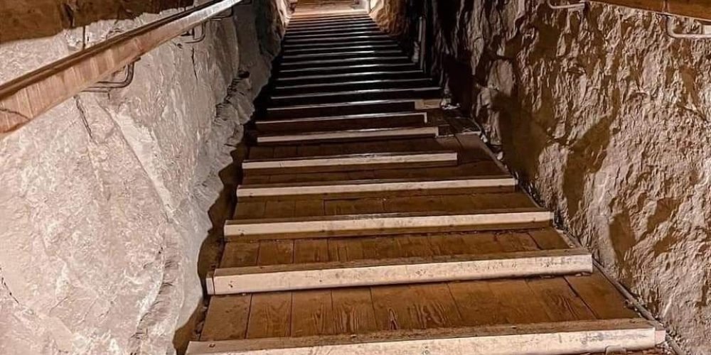 A long, narrow, and ascending wooden staircase located inside the Great Pyramid. The stairs have metal edges and are flanked by low wooden handrails on both sides. The walls are made of rough, irregular limestone blocks, illuminated by warm artificial lighting hidden along the sides, creating a tunnel-like perspective that leads toward a brightly lit opening at the top.