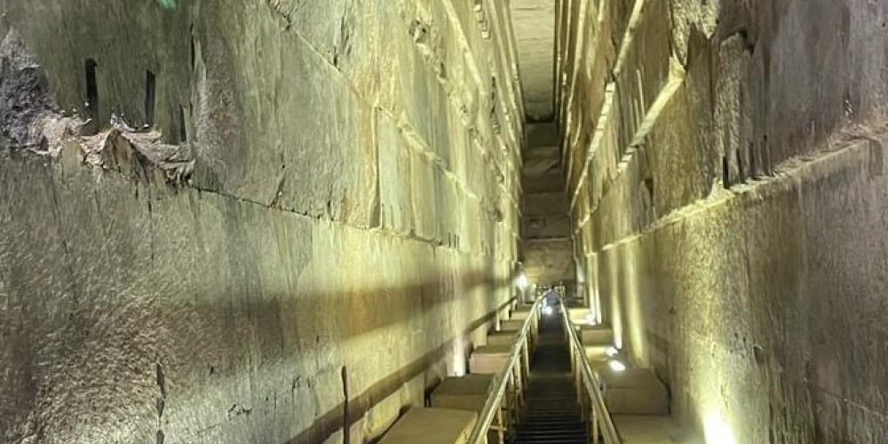 A close-up selfie of a smiling man and woman at the bottom of the frame, positioned within the narrow, soaring Grand Gallery inside the Great Pyramid. The architectural space behind them features high, corbelled limestone walls that taper toward the ceiling. A steep wooden staircase with metal safety treads and a handrail extends deep into the background, illuminated by bright white artificial floor lights that highlight the ancient stone textures.