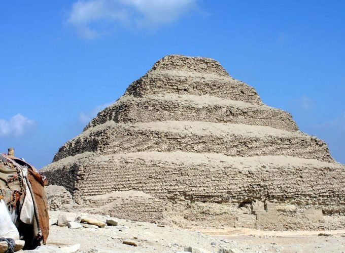 A wide, clear outdoor shot featuring a white camel sitting on the sandy ground in the bottom-left foreground, wearing a colorful decorative saddle. Behind it, the iconic Step Pyramid of Djoser at Saqqara dominates the center, showing its six receding stone tiers. The ground is a light-colored desert floor with scattered rocks, and the background is a bright blue sky with a few soft white clouds, captured during a Saqqara VIP tour
