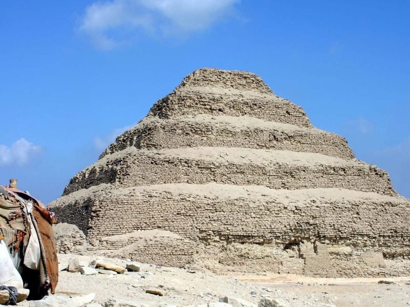 A wide, clear outdoor shot featuring a white camel sitting on the sandy ground in the bottom-left foreground, wearing a colorful decorative saddle. Behind it, the iconic Step Pyramid of Djoser at Saqqara dominates the center, showing its six receding stone tiers. The ground is a light-colored desert floor with scattered rocks, and the background is a bright blue sky with a few soft white clouds, captured during a Saqqara VIP tour