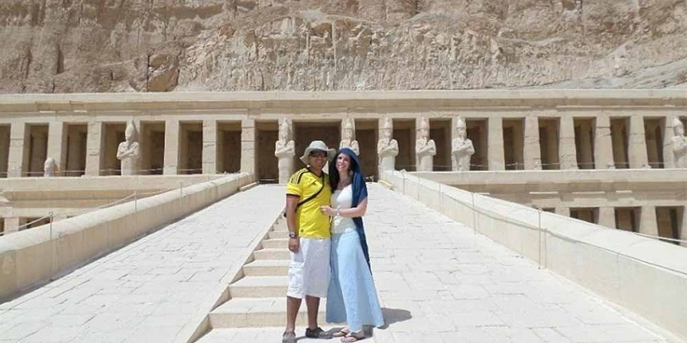 Um casal sorridente posa na rampa de pedra central do Templo de Hatchepsut, em Luxor. O homem veste uma t-shirt amarela, calções brancos e chapéu. A mulher veste uma blusa branca, uma saia azul comprida e um lenço azul na cabeça. Ao fundo, avistam-se as colunas do templo com estátuas de Osíris e os imponentes penhascos de calcário. Um dos pontos altos de uma viagem ao Egito.