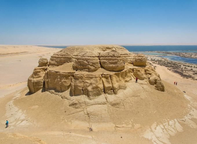 Vue aérienne d'une immense formation rocheuse naturelle de calcaire beige de forme circulaire et aplatie au sommet, présentant des parois érodées verticalement. Elle se dresse seule au milieu d'un désert de sable clair, bordée à l'horizon par l'eau bleue d'un lac et un ciel azur sans nuages lors d'un voyage vers le Fayoum depuis le Caire