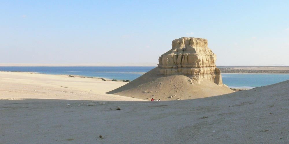 Une vaste colline naturelle de calcaire ocre à la forme arrondie et au sommet plat, sculptée de rainures verticales par l'érosion, se dressant seule dans un désert de sable clair. À droite, on aperçoit une étendue d'eau bleue et des zones de végétation basse près du rivage lors d'une excursion vers le Fayoum depuis le Caire