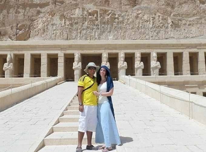 Un couple souriant pose sur la rampe centrale en pierre du temple d'Hatchepsout à Louxor. L'homme porte un t-shirt jaune, un short blanc et un chapeau. La femme porte un haut blanc, une jupe longue bleue et un voile bleu sur la tête. En arrière-plan, on voit les colonnes du temple avec des statues d'Osiris et les falaises de calcaire massives. Une étape phare d'un Voyage en Egypte