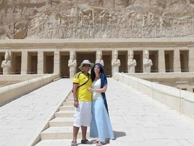 Un couple souriant pose sur la rampe centrale en pierre du temple d'Hatchepsout à Louxor. L'homme porte un t-shirt jaune, un short blanc et un chapeau. La femme porte un haut blanc, une jupe longue bleue et un voile bleu sur la tête. En arrière-plan, on voit les colonnes du temple avec des statues d'Osiris et les falaises de calcaire massives. Une étape phare d'un Voyage en Egypte