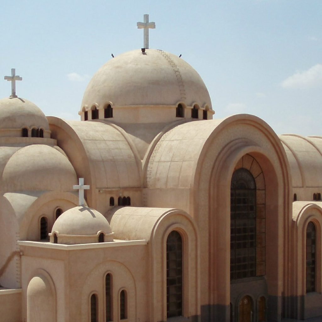 A close-up architectural view of the Saint Bishoy Monastery featuring several large, tan-colored domes topped with white Christian crosses. The building showcases traditional Coptic design with arched entryways and small rectangular windows under a clear sky during a Wadi El Natrun Tour. The smooth, sandy texture of the stone exterior reflects the peaceful desert environment characteristic of this historic monastic site