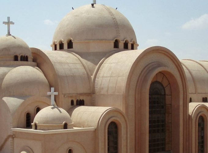 A close-up architectural view of the Saint Bishoy Monastery featuring several large, tan-colored domes topped with white Christian crosses. The building showcases traditional Coptic design with arched entryways and small rectangular windows under a clear sky during a Wadi El Natrun Tour. The smooth, sandy texture of the stone exterior reflects the peaceful desert environment characteristic of this historic monastic site