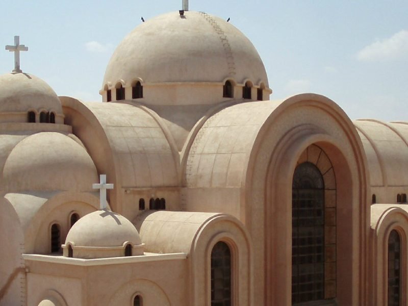 A close-up architectural view of the Saint Bishoy Monastery featuring several large, tan-colored domes topped with white Christian crosses. The building showcases traditional Coptic design with arched entryways and small rectangular windows under a clear sky during a Wadi El Natrun Tour. The smooth, sandy texture of the stone exterior reflects the peaceful desert environment characteristic of this historic monastic site