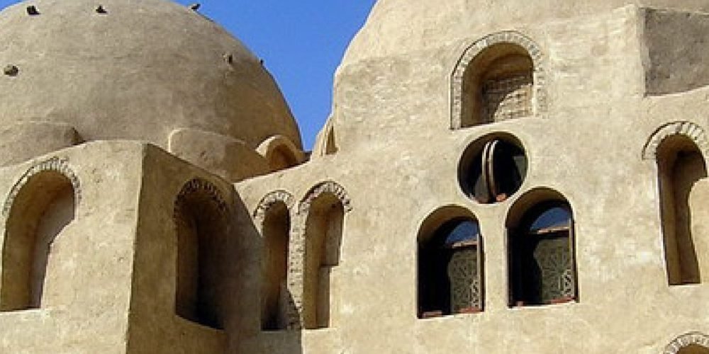 A close-up view of the ancient stone walls and rounded desert domes of a historic monastery during a Wadi El Natrun Tour. The structure features multiple arched windows with brick detailing and small circular openings set against a clear blue sky. The weathered tan limestone texture of the monastery highlights the traditional Coptic architectural style found in the Egyptian desert