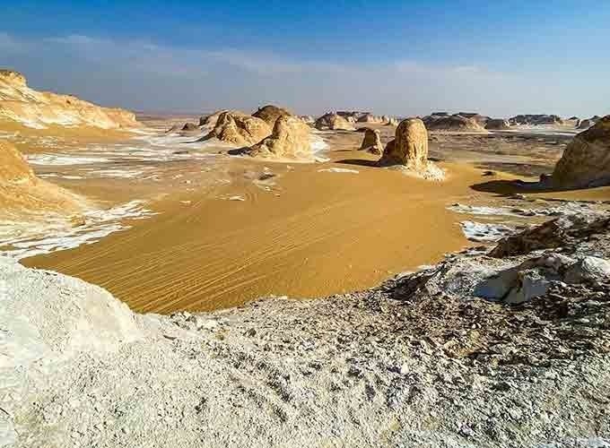 A wide landscape view of the White desert featuring unique chalk rock formations and towering white mushrooms rising from golden sands. The terrain shows a mix of white limestone surfaces and orange desert dunes under a vast, clear blue sky with light hazy horizons. In the foreground, a rocky white plateau overlooks a valley of natural geological sculptures and scattered hills in the distance
