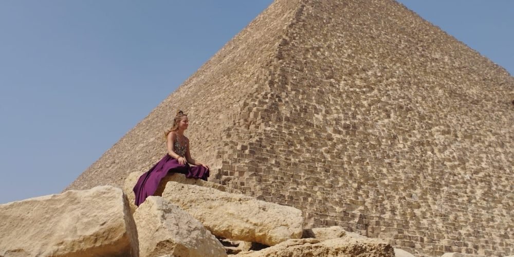 journée au musée égyptien des pyramides de Gizeh1 Une femme portant une longue robe violette est assise sur de gros blocs de calcaire beige clair au premier plan. Derrière elle s'élève la paroi immense et inclinée d'une grande pyramide faite de milliers de pierres empilées, sous un ciel bleu uni lors d'une journée au musée égyptien des pyramides de Gizeh