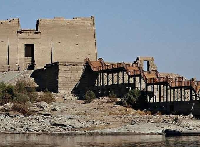 Une vue depuis l'eau du temple de Kalabsha, un grand édifice de pierre aux murs massifs et plats. Un long ponton en bois avec des escaliers en zigzag mène du bord du fleuve jusqu'à l'entrée du monument, sur une rive rocheuse parsemée de petits buissons verts lors d'une journée au musée nubien et temple de Kalabsha