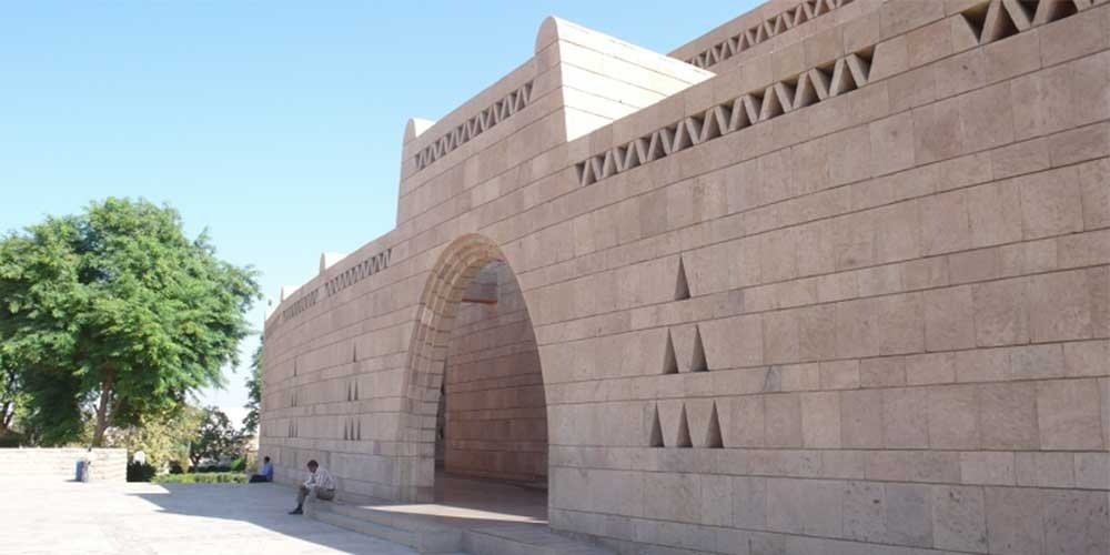 Une vue de la façade du musée nubien construite en blocs de pierre ocre avec des motifs triangulaires sculptés. Une grande arche centrale mène à l'intérieur, tandis qu'un homme est assis sur un rebord à l'ombre à gauche, sous un ciel bleu lors d'une journée au musée nubien et temple de Kalabsha
