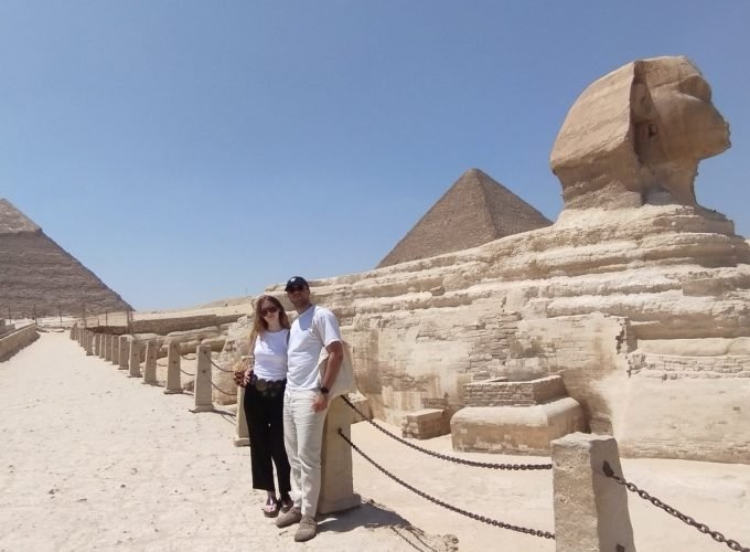 Un jeune couple pose debout sur un chemin de sable à côté du Grand Sphinx. La femme porte un t-shirt blanc, un pantalon noir et des lunettes de soleil. L'homme porte un t-shirt blanc, un pantalon clair et une casquette noire. En arrière-plan, on voit clairement deux grandes pyramides sous un ciel bleu intense sans nuages. Des poteaux avec des chaînes délimitent le passage lors de cette journée aux pyramides de Gizeh