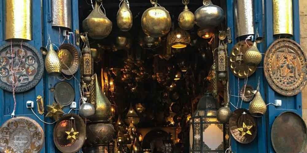 Loja tradicional de lanternas de metal no Cairo, exibindo candeeiros suspensos de latão, pratos gravados e artigos decorativos feitos à mão. Luzes douradas e aconchegantes preenchem a estreita entrada da loja, criando uma autêntica atmosfera oriental. Uma cena vibrante de um mercado local, vivida durante uma viagem ao Cairo.