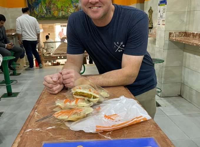 Un homme souriant en t-shirt bleu marine et lunettes de soleil sur la tête s'appuie sur un comptoir en marbre brun. Devant lui posent trois sandwichs égyptiens (Falafel) emballés dans du plastique. Sur le comptoir, on voit une pancarte bleue "EXPLORE EGYPT TOURS". L'arrière-plan montre l'intérieur d'un restaurant avec des murs orange, des tabourets verts et une fresque colorée illustrant la vie égyptienne. Une scène authentique de visite gastronomique