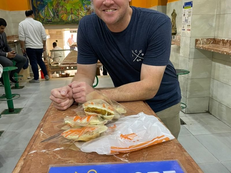 Un homme souriant en t-shirt bleu marine et lunettes de soleil sur la tête s'appuie sur un comptoir en marbre brun. Devant lui posent trois sandwichs égyptiens (Falafel) emballés dans du plastique. Sur le comptoir, on voit une pancarte bleue 