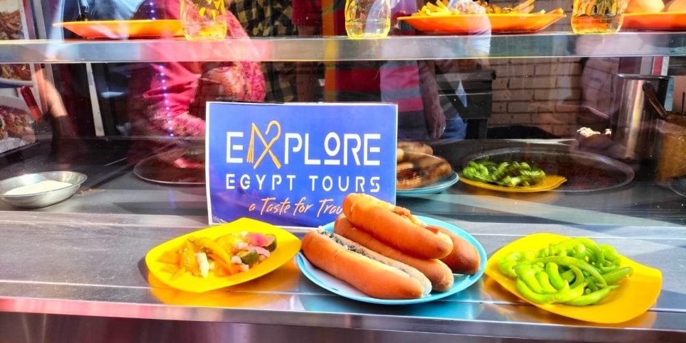 visite gastronomique3 Une vitrine de restaurant en acier inoxydable présentant plusieurs plats : au centre, une assiette bleue avec trois sandwichs allongés, flanquée de deux assiettes jaunes contenant des piments verts marinés et des légumes variés. Derrière la vitre, on voit un panneau bleu "EXPLORE EGYPT TOURS", des bouteilles d'huile dorées et le reflet de personnes qui regardent. Une étape gourmande d'une visite gastronomique.