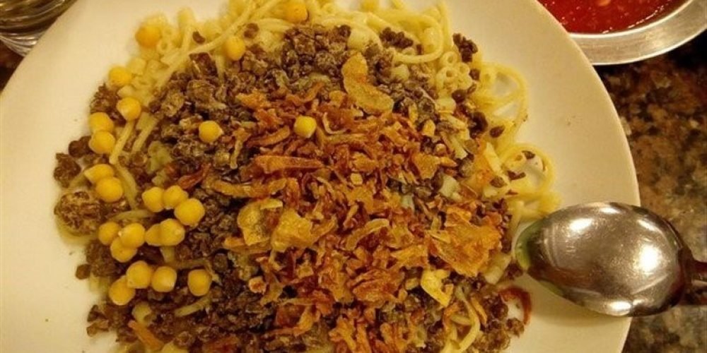 visite gastronomique4 Une vue panoramique d'une plage de sable fin bordée par les eaux cristallines et peu profondes de la mer Rouge. Le rivage est paré de nombreux parasols en paille et de palmiers hauts et fins sous un ciel bleu éclatant. On aperçoit des bâtiments de resort blancs en arrière-plan, créant un cadre idéal pour une pause lors d'une visite gastronomique