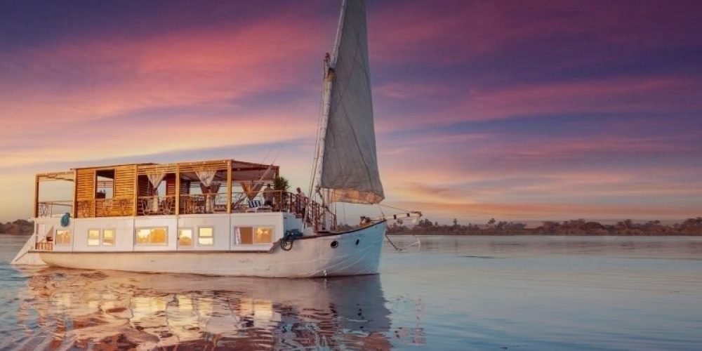 A luxury white Dahabyia sailing boat with a large wooden deck gliding on the Nile River during a vibrant sunset.