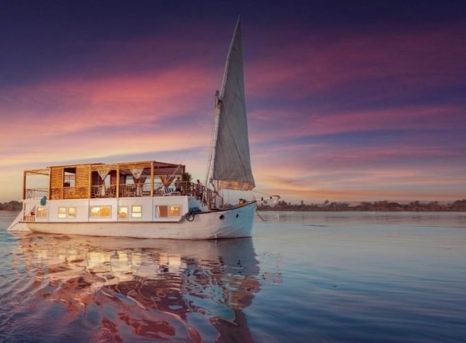 A luxury white Dahabyia sailing boat with a large wooden deck gliding on the Nile River during a vibrant sunset.