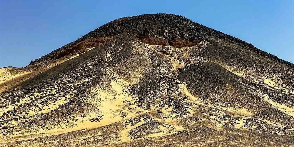 A majestic black volcanic hill rising from the golden sands of the Egyptian desert under a clear blue sky
